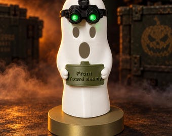 Könnte beinhalten: Eine weiße Geisterfigur mit einer schwarzen Nachtsichtbrille mit leuchtend grünen Linsen. Der Geist hält ein olivgrünes rechteckiges Schild mit dem geprägten Text Front Toward Enemy. Er steht auf einem runden, goldenen Sockel.