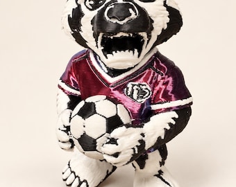 Könnte beinhalten: Eine 3D-gedruckte Dachsfigur mit schwarz-weißem Fell, die ein metallisch violett-blaues Fußballtrikot mit einem kreisförmigen Emblem auf der Brust trägt. Der Dachs hat ein offenes Maul mit sichtbaren Zähnen und hält einen Fußball.