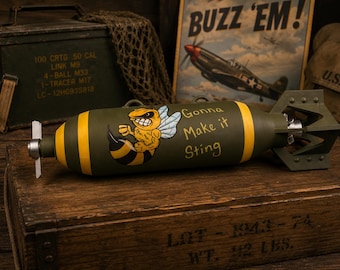 Könnte beinhalten: Eine olivgrüne, zylindrische Bombenattrappe mit gelb gestreifter Nase und Heck, einer Hornissen-Grafik und dem Text "Gonna Make it Sting" in gelber Schrift. Das Objekt verfügt über einen weißen Propeller an der Vorderseite und Metallflossen.