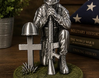 May include: A silver-toned figurine depicting a soldier kneeling in prayer with hands clasped over the barrel of an upright rifle. Beside the soldier stands a cross topped with a helmet, all set upon a circular base with a textured green surface.