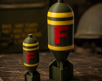 Könnte beinhalten: Zwei olivgrüne, bombenförmige Objekte in verschiedenen Größen, jeweils mit zwei horizontalen gelben Streifen und einem markanten roten Buchstaben F an der Seite. Die Objekte haben eine abgerundete Oberseite mit einer kleinen Öse und eine Leitwerkbasis.