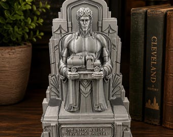 Könnte beinhalten: Eine graue 3D-gedruckte Skulptur einer muskulösen Figur, die auf einem Thron sitzt und ein kleines LKW-Modell hält. Der Sockel trägt den geprägten Text 'Industry Builds Cleveland Endures'. Die Figur hat geflochtenes Haar und einen architektonischen Hintergrund.