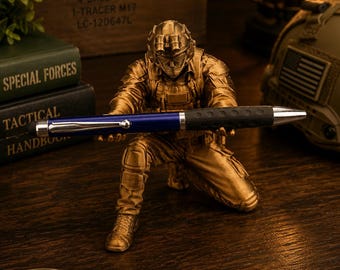 Könnte beinhalten: Eine goldfarbene Soldatenfigur in kniender Position, die einen blauen und schwarzen Kugelschreiber hält. Der Soldat trägt taktische Ausrüstung, einschließlich eines Helms mit Schutzbrille. Der Stift hat einen silbernen Clip und einen schwarzen Griff.