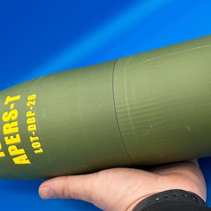 May include: Olive green artillery shell with yellow text reading "105 M APERS-T LOT-DBP-26" and a yellow tip. A red band wraps around the middle of the shell. The shell is resting on a blue surface.