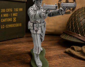 Könnte beinhalten: Eine silberfarbene Soldatenfigur auf einem grünen Sockel, die einen Helm und eine Uniform trägt und zwei Handfeuerwaffen in einer Schusshaltung nach vorne hält. Die Figur zeigt detaillierte Gesichtszüge, taktische Ausrüstung und Schnürstiefel.