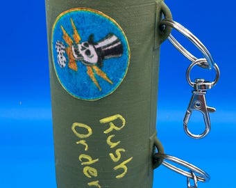 Könnte beinhalten: Ein grünes, bombenförmiges Novelty-Item mit Propeller, Flossen und einem runden Aufnäher mit Totenkopf und Blitzen. Der Text "Rush Order" steht auf der Seite, und es sind zwei silberne Karabinerhaken angebracht.