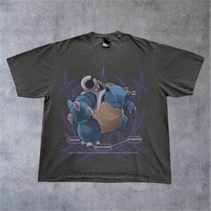 Puede incluir: Camiseta gris oscuro con un personaje de dibujos animados Blastoise azul y beige. El diseño incluye las palabras "WATER" y "STARTER" sobre un fondo morado con forma de telaraña.