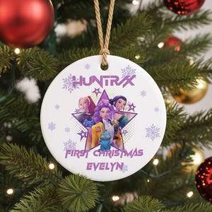 May include: A white ceramic Christmas ornament with a star-shaped graphic featuring three cartoon characters. The ornament has the text "HUNTRAX" and "FIRST CHRISTMAS EVELYN" in purple. The ornament hangs from a natural twine string.