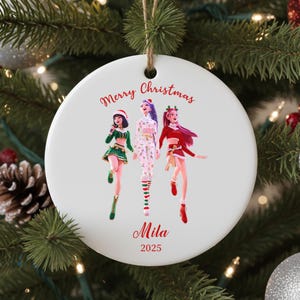 Op de afbeelding: Ronde witte keramische kerstversiering met een rood touwtje om op te hangen. De versiering toont drie cartoonfiguren in feestelijke outfits, de woorden "Merry Christmas" en de naam "Mila" in het rood, en het jaar 2025.
