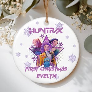 May include: White ceramic Christmas ornament featuring a graphic of three animated women inside a purple star. The ornament has the text "HUNTRAX" at the top and "FIRST CHRISTMAS EVELYN" at the bottom, with snowflake accents.