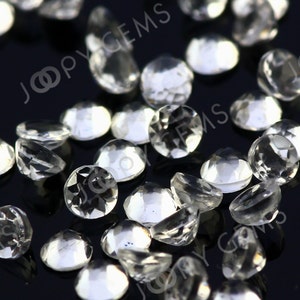May include: A collection of clear, faceted gemstones scattered on a dark surface. The gems are round and have multiple facets that reflect light, creating a sparkling effect. The image is well-lit, highlighting the clarity and brilliance of the stones. The text "JOOPY GEMS" is visible in the background.