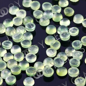 Puede incluir: Una colecci&oacute;n de peque&ntilde;as gemas redondas de color verde p&aacute;lido con una superficie pulida y abovedada. Las gemas est&aacute;n dispersas sobre una superficie oscura, mostrando su cualidad transl&uacute;cida y sutiles variaciones de color. Ideales para joyer&iacute;a.