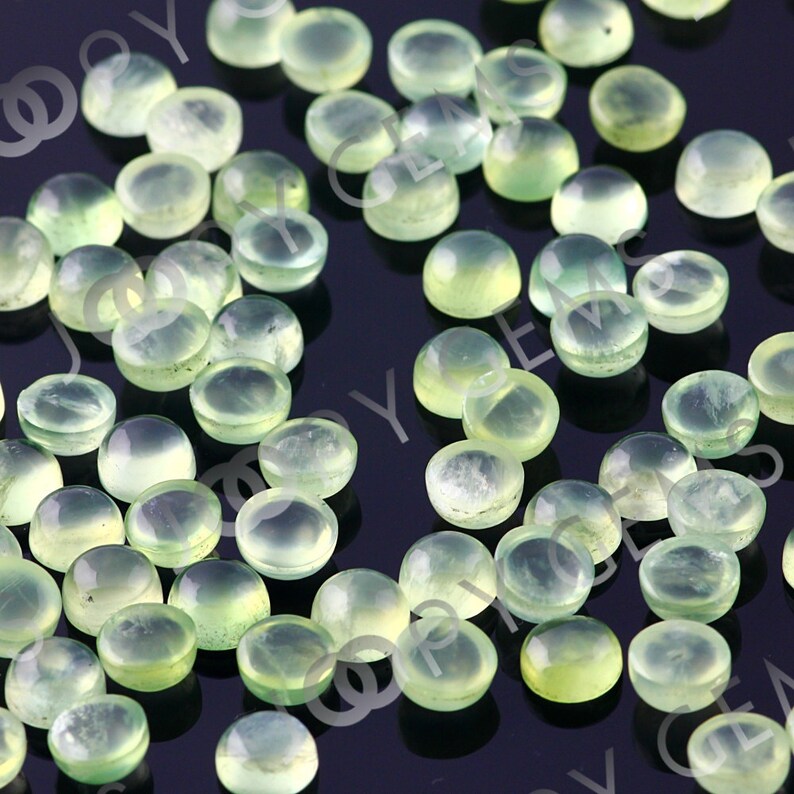 Puede incluir: Una colecci&oacute;n de peque&ntilde;as gemas redondas de color verde p&aacute;lido con una superficie pulida y abovedada. Las gemas est&aacute;n dispersas sobre una superficie oscura, mostrando su cualidad transl&uacute;cida y sutiles variaciones de color. Ideales para joyer&iacute;a.