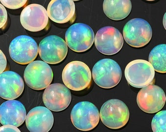 Può includere: Una collezione di cabochon di opale rotondi e iridescenti in varie tonalità di blu, verde e rosa. I cabochon sono sparsi su una superficie nera.