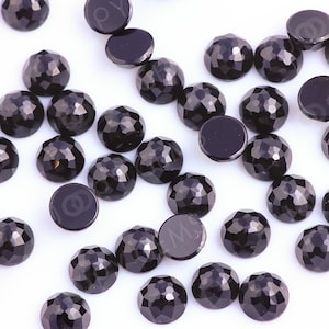 Puede incluir: Una colección de piedras preciosas redondas facetadas en cabujón de color negro. Las piedras están esparcidas sobre una superficie blanca.