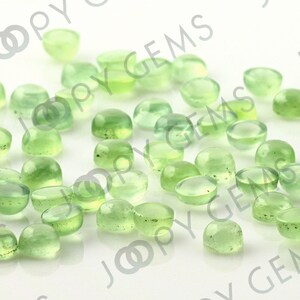 Puede incluir: Una colecci&oacute;n de piedras preciosas cabuj&oacute;n transl&uacute;cidas de color verde claro. Las piedras preciosas son redondas con una base plana y una parte superior abovedada. Las gemas est&aacute;n dispersas sobre una superficie blanca, con el texto "JOOPY GEMS" visible en el fondo.