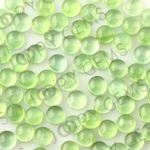 Puede incluir: Una colecci&oacute;n de piedras preciosas redondas y transl&uacute;cidas de color verde claro. Las piedras est&aacute;n dispersas sobre una superficie blanca, con diferentes tonos de verde. El efecto general es una exhibici&oacute;n suave y luminosa de piedras naturales.