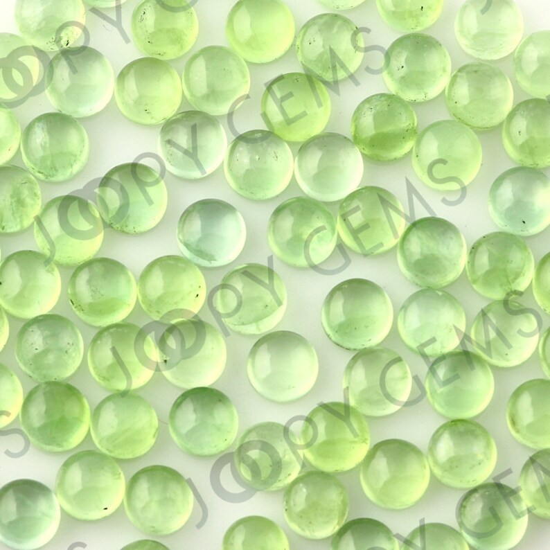 Puede incluir: Una colecci&oacute;n de piedras preciosas redondas y transl&uacute;cidas de color verde claro. Las piedras est&aacute;n dispersas sobre una superficie blanca, con diferentes tonos de verde. El efecto general es una exhibici&oacute;n suave y luminosa de piedras naturales.