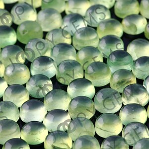 Puede incluir: Primer plano de numerosas piedras preciosas redondas y transl&uacute;cidas en tonos verdes y verde azulado. Las piedras talladas en cabuj&oacute;n tienen una superficie pulida. La imagen tiene un enfoque suave, destacando los colores y texturas naturales.
