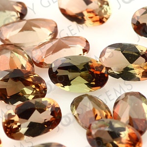 May include: A collection of oval-cut gemstones in shades of brown, amber, and olive green. The faceted stones are arranged closely together, showcasing their polished surfaces and internal clarity. The gemstones appear to be of high quality, with a warm, inviting color palette.