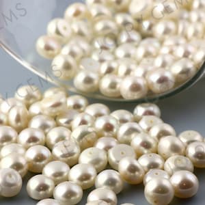 May include: A close-up of numerous creamy white pearls, some with a subtle sheen, cascading from a clear glass vessel. The pearls are spherical and polished, suitable for jewellery or crafts. The lighting accentuates the pearls' surface detail.