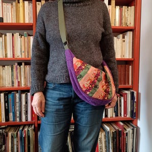 Peut inclure: Un sac à bandoulière violet avec un motif patchwork dans les tons de rose, vert et orange. Le sac a une forme incurvée et une sangle verte. La personne porte un pull gris et un jean bleu.
