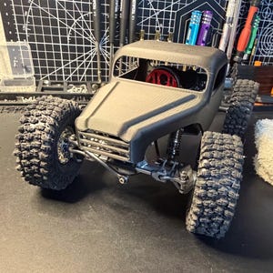 May include: A gray, miniature off-road truck model with large, textured tires. The truck has a detailed chassis and a red component visible inside. The model is on a dark surface, with tools and a grid mat in the background.