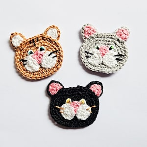 May include: Three crocheted cat faces, one orange, one gray, and one black. Each face has pink ears and a white nose.