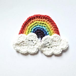 May include: A crocheted rainbow with white clouds on either side. The rainbow is made of red, orange, yellow, green, blue, indigo, and violet yarn. The clouds are made of white yarn.