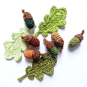 Op de afbeelding: Een verzameling gehaakte eikels en eikenbladeren in tinten groen, bruin en oranje. De eikels zijn klein en gedetailleerd, met een dop en een noot. De bladeren zijn realistisch en hebben een textuur.