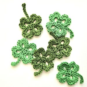 以下が含まれることがあります： 白い背景に、それぞれ異なるデザインの緑色のクロシェ編みされた四つ葉のクローバーが6つあります。