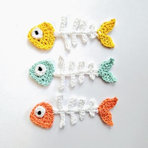 Può includere: Tre disegni di lisca di pesce all'uncinetto in giallo, verde acqua e arancione. Ogni pesce ha una struttura ossea bianca, un occhio a contrasto e una coda abbinata. I pesci sono fatti a mano e sembrano decorativi.