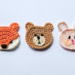 Puede incluir: Tres caras de animales de ganchillo: un zorro naranja y blanco, un oso marrón y un conejito rosa. Cada cara es redonda con detalles bordados, ideal para manualidades o para adornar artículos infantiles. El zorro tiene ojos negros y cara blanca.