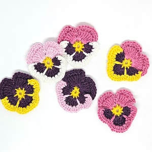 Può includere: Sei fiori di viola del pensiero all'uncinetto in tonalità di rosa, viola e giallo. I fiori sono tutti a forma di cuore e hanno un cerchio giallo centrale.