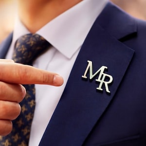Peut inclure: Épinglette argentée avec les lettres « MR » fixée à une veste de costume bleu marine. La veste est portée sur une chemise blanche à col et une cravate à motifs. Un doigt pointe vers l'épinglette.