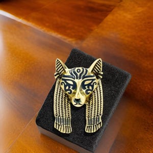 Könnte beinhalten: Eine goldfarbene Brosche mit einem schwarzen und goldenen Design eines Tierkopfes mit einem stilisierten Gesicht. Die Brosche befindet sich auf einem schwarzen Samt-Hintergrund.