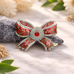 Peut inclure: Broche décorative en forme de nœud, avec une finition émail rouge et des accents dorés. Le nœud est orné de perles turquoise, de cristaux transparents et d'une pierre rouge centrale. Des détails floraux verts complètent le design.