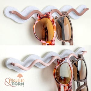 May include: A wall-mounted sunglasses holder with a wavy, light gray felt base and curved, light brown arms. Two pairs of sunglasses hang from the holder, one with a tortoiseshell pattern and the other with a wood grain design. The logo "Flourish & Form" is visible.