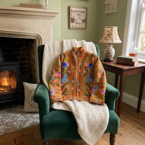 Puede incluir: Chaqueta bordada de color mostaza con diseños florales y de elefantes, exhibida en un sillón de terciopelo verde. La chaqueta presenta detalles en rosa, azul y naranja. Una manta de punto color crema está sobre la silla.