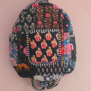 May include: A black quilted backpack with a patchwork design featuring floral and geometric patterns in shades of pink, blue, and white. The backpack has a small pocket on the front and adjustable straps.