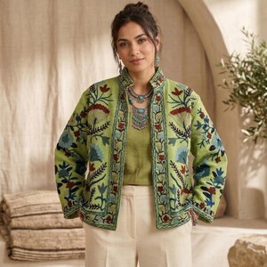 Op de afbeelding: Lichtgroene geborduurde jas met een bloemenmotief in rood, blauw en groen. De jas wordt gedragen over een bijpassende groene top en gecombineerd met crèmekleurige broek. De jas heeft een opstaande kraag en ingewikkelde borduursels.
