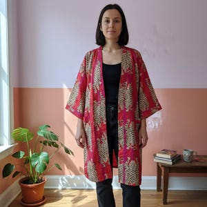 May include: A red kimono-style robe with a tiger stripe pattern. The robe is open in the front and worn over a black top and black jeans. The background is a two-tone wall with a pink and peach color scheme.