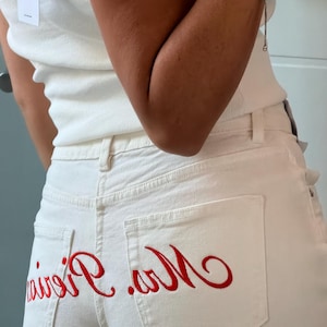 Puede incluir: Vaqueros blancos con texto rojo bordado en los bolsillos traseros. Los vaqueros tienen un diseño clásico de cinco bolsillos y cierre de botón y cremallera. Una camiseta sin mangas blanca es visible en la parte superior de la imagen.