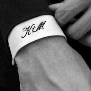 May include: A white shirt cuff featuring black embroidered cursive lettering that reads K&M. The cuff is visible against a dark sleeve, showcasing the detailed stitching of the monogram on the crisp white fabric of the shirt sleeve.