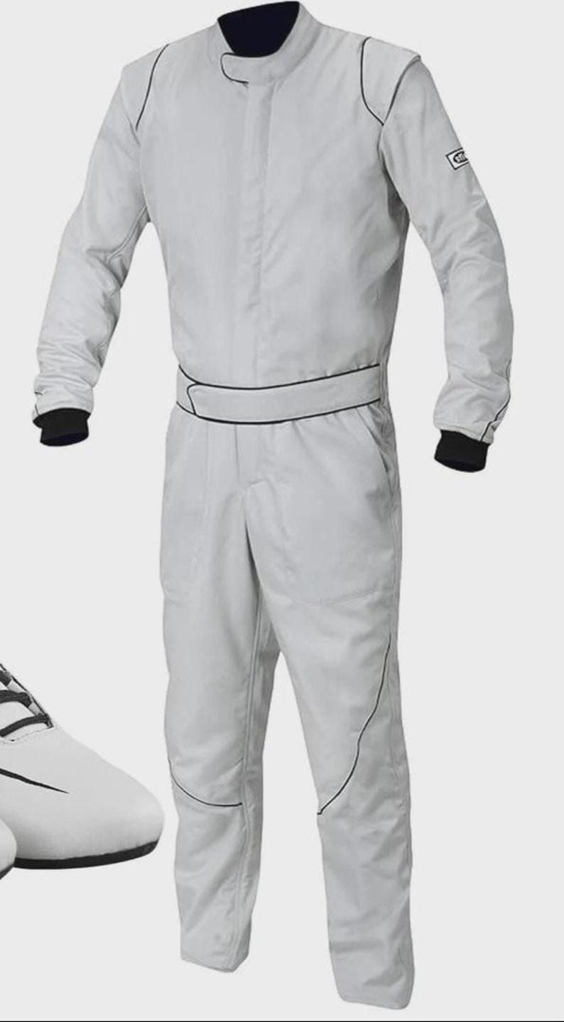 May include: Light gray racing suit with black trim and cuffs. The one-piece suit features a stand-up collar and a black belt detail. The suit is shown with white racing shoes. This racing suit is designed for motorsports.