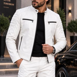 May include: A white leather jacket featuring a mandarin collar with snap buttons, quilted shoulder panels, and black zippered pockets on the chest and waist. The jacket is worn over a black shirt and paired with matching white trousers.