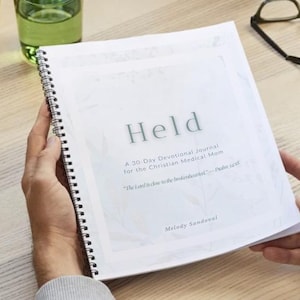以下が含まれることがあります： Held, A 30-Day Devotional Journal for the Christian Medical Momというタイトルのスパイラル綴じのデボーションジャーナル。淡い植物の背景、サブタイトルThe Lord is close to the brokenhearted—Psalm 34:18、著者Melody Sandovalの記載がある。