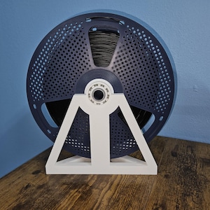 May include: A dark blue filament spool holder with a white support structure. The spool is perforated with small holes and is mounted on a white, triangular base. The holder is designed to keep the filament organized.
