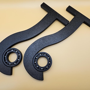 Puede incluir: Dos objetos metálicos negros y curvos con un acabado texturizado, cada uno con un rodamiento circular con bolas de acero plateadas. Los objetos tienen un asa en forma de T. El fondo es de color beige claro.