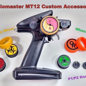 May include: Radiomaster MT12 custom accessories for RC vehicles.  The image shows a black radio transmitter with a pink and gold yin-yang design on the wheel.  There are also various colored knobs, steering wheel grips, and wheel grips.